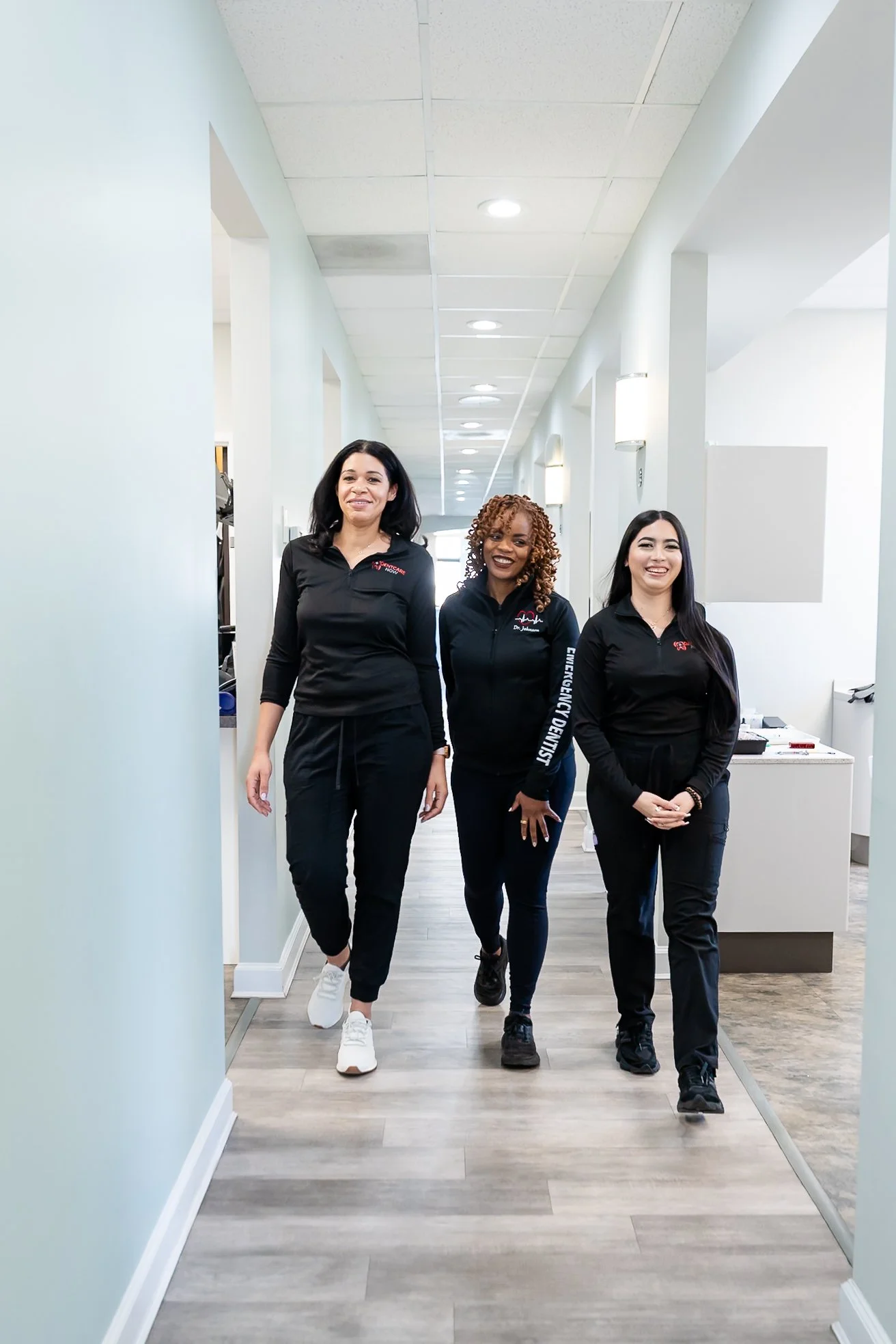 Three Dentcare Now team members walking down a modern dental office hallway in black uniforms